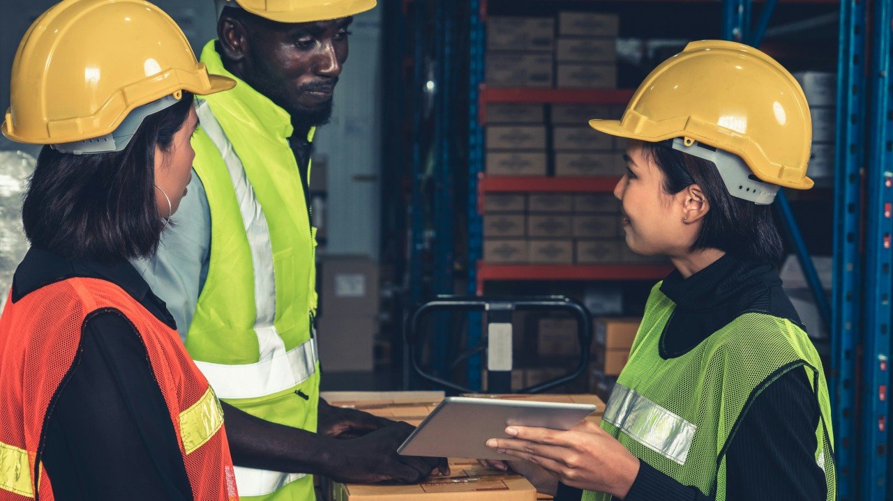 Three warehouse workers in high-visibility vests and hard hats reviewing a tablet together, the kind of operational collaboration that delivers for customers