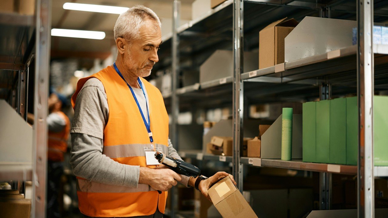 A warehouse worker in a safety vest scanning a package on a stocked shelf, where the real work happens